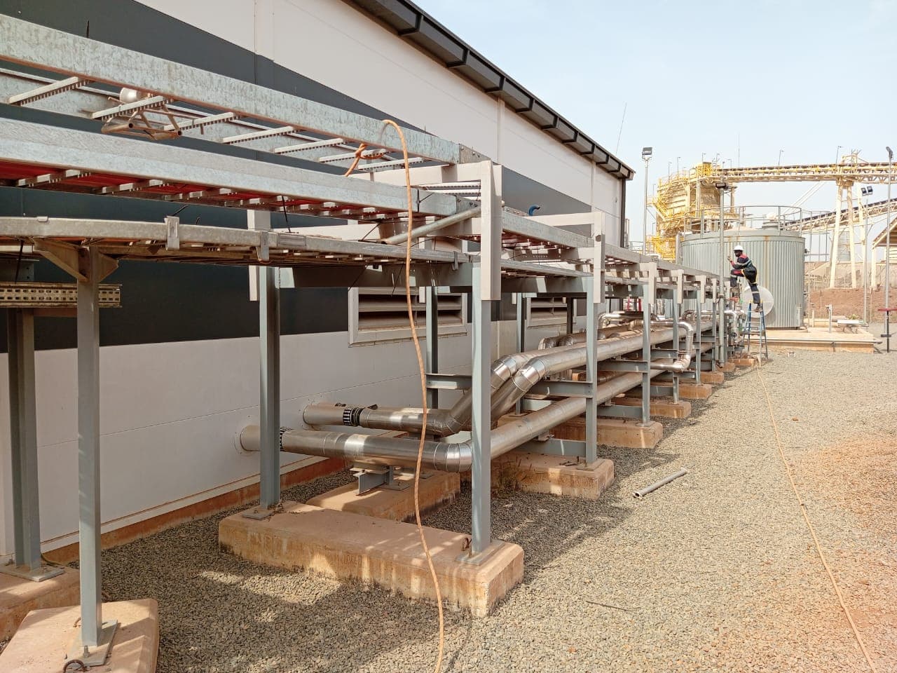 Chemins de câbles galvanisés le long d’un bâtiment industriel sur site minier, technicien intervenant au pied d’une cuve de process